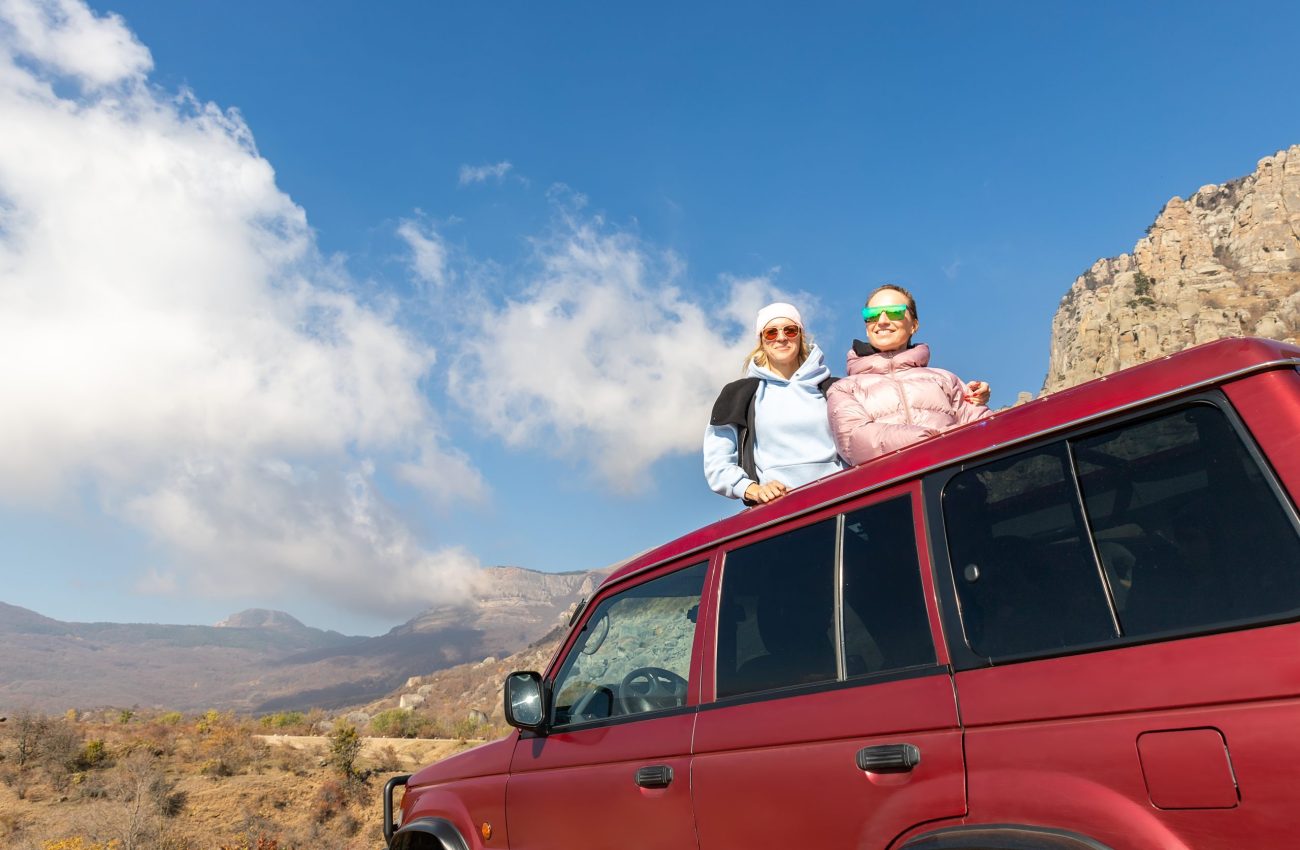 Scenic view two young adult woman friends enjoy having fun sit on car roof at adventure suv offroad mountain trip against blue sky on sunny autumn day. Female party nature trial journey concept.